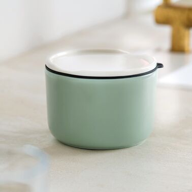 Runde Lunchbox aus mintgrünem Premium Porzellan mit weißem Deckel und schwarzer Silikondichtung steht auf einer hellen, glatten Oberfläche. Das minimalistische Design wirkt modern und hochwertig. Im Hintergrund leicht verschwommene Küchendetails erkennbar.