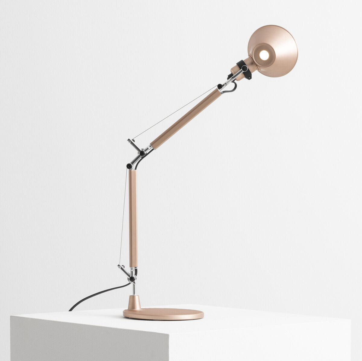 Artemide Tolomeo Micro Tischleuchte in Kupferoptik auf einem weißen Podest. Modernes und minimalistisches Design mit verstellbarem Arm und rundem Lampenkopf.