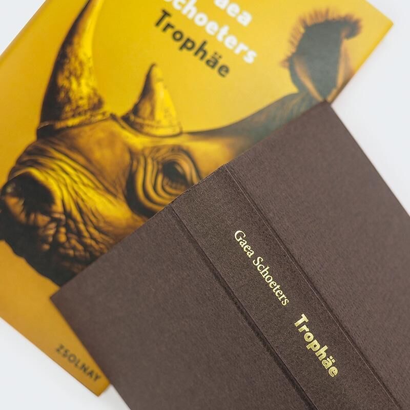 Das Bild zeigt das Buch 'Trophäe' von Gaea Schoeters. Der Buchumschlag ist in Gelb- und Brauntönen gestaltet und zeigt die Abbildung eines Nashorns. Das Buch liegt auf einer flachen Oberfläche neben einer weiteren, teilweise sichtbaren Broschüre oder Buch.