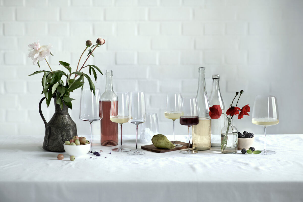 Ein stilvoll gedeckter Tisch mit mehreren Weingläsern des Sommelier-Bundles »Vervino«, gefüllt mit Rotwein und Weißwein. Dazu verschiedene Weinflaschen, eine Karaffe, Blumen in einer Vase, grüne und rote Trauben, und eine Birne.