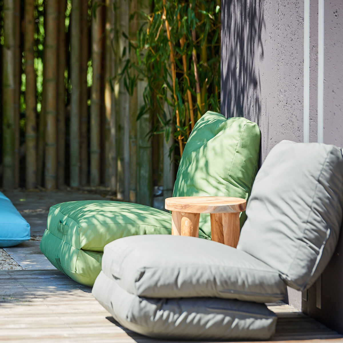 Gartenmatratze »Sheffield« auf einer Holzterrasse im Freien, umgeben von grünen Pflanzen. Es gibt grüne und blaue Kissen und einen kleinen Holztisch.