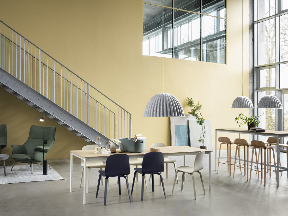 Ein modern eingerichtetes Zimmer mit einem großen Tisch, grauen Aufbewahrungskörben, Stühlen in verschiedenen Farben und minimalistischen Dekorationen. Eine metallene Treppe führt zu einem oberen Bereich, und große Fenster lassen viel Licht herein.