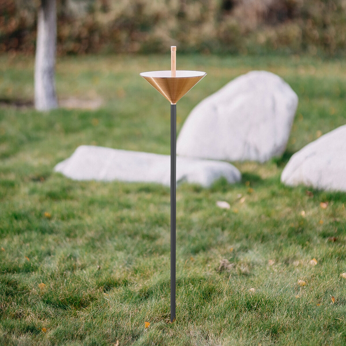 Der Regenmesser Kupfer der ZEIT-Sonderedition von Fischer Manufaktur steht auf einer Rasenfläche. Er besteht aus einem konischen Kupferkegel auf einem langen, pulverbeschichteten Stab. Im Hintergrund sind große, helle Steine und Bäume im unscharfen Gartenbereich sichtbar.