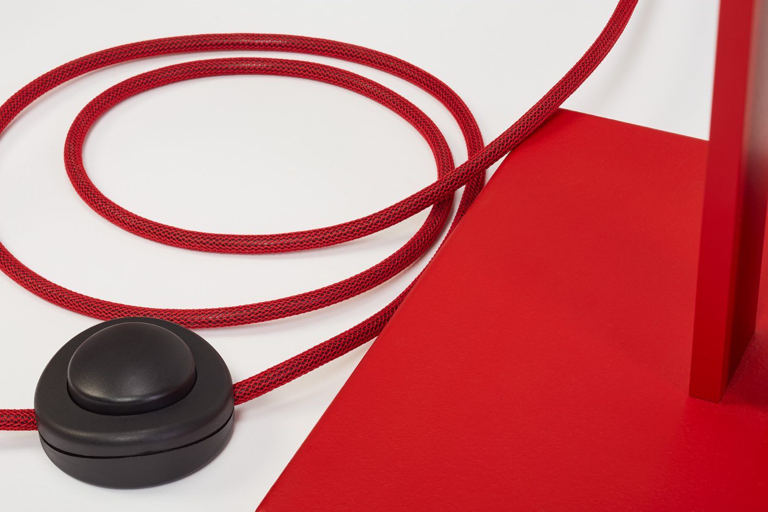 Detailaufnahme des roten, textilummantelten Stromkabels der Stehleuchte »schliephacke« von mawa design auf weißer und roter Oberfläche. Sichtbar ist zudem ein großer, runder, schwarzer Fußschalter sowie der kantige, rote Lampenfuß. Hochwertige Materialien und modernes Design dominieren das Bild.