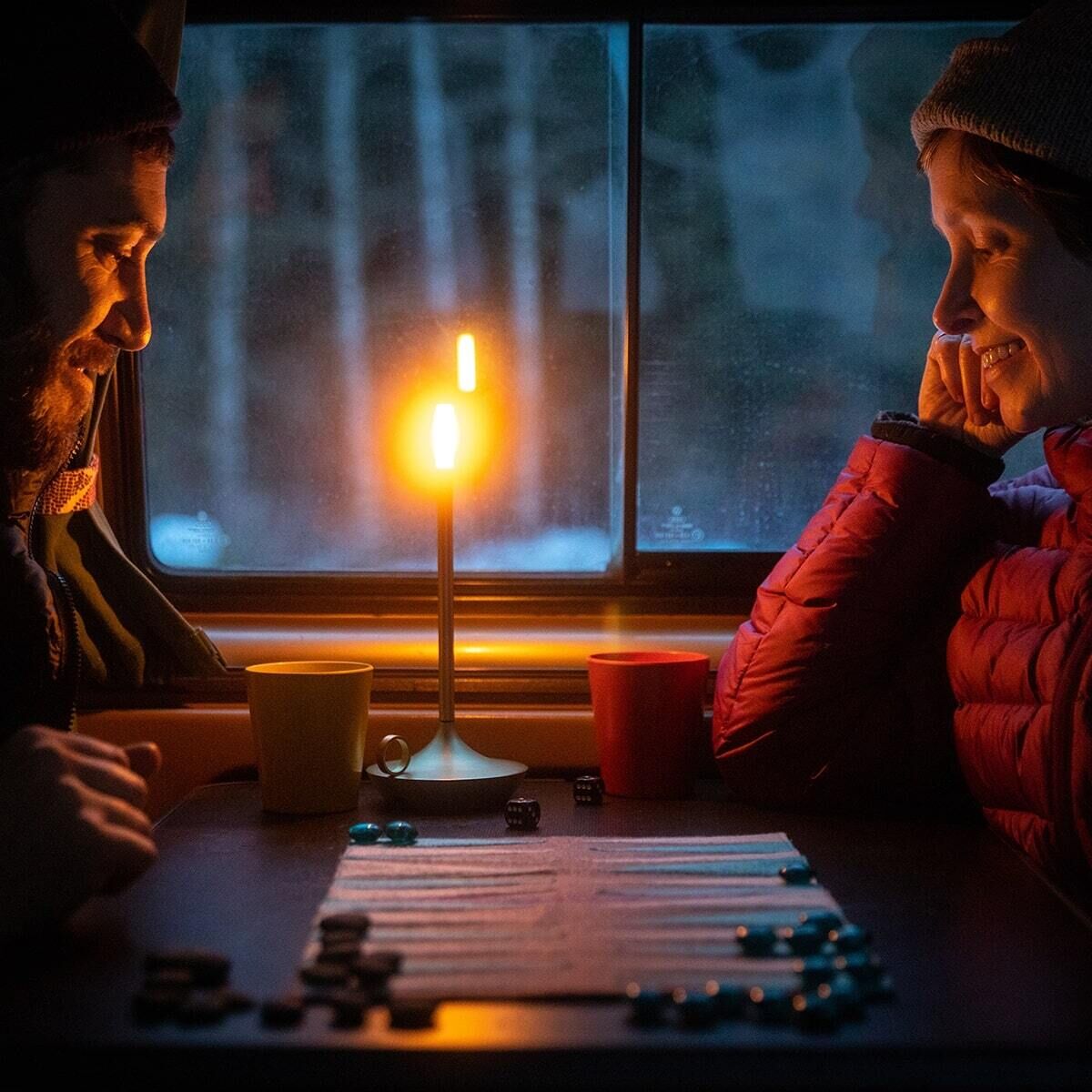 Zwei Personen genießen die behagliche Atmosphäre mit der Wick Tischlampe, die warmes Kerzenlicht ausstrahlt, während sie ein Brettspiel spielen. Im Hintergrund sieht man ein Fenster mit einer winterlichen Landschaft.