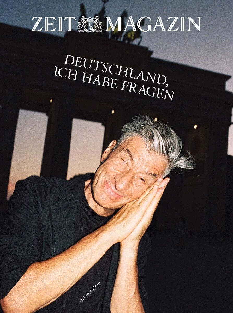 Titelbild des ZEIT Magazins 37/2026. Großaufnahme eines lachenden Mannes mit grauen Haaren und schwarzen Kleidern, der seine Hände wie zum Schlafen an die Wange legt. Im Hintergrund das beleuchtete Brandenburger Tor bei Abenddämmerung. Text: 'DEUTSCHLAND, ICH HABE FRAGEN.'