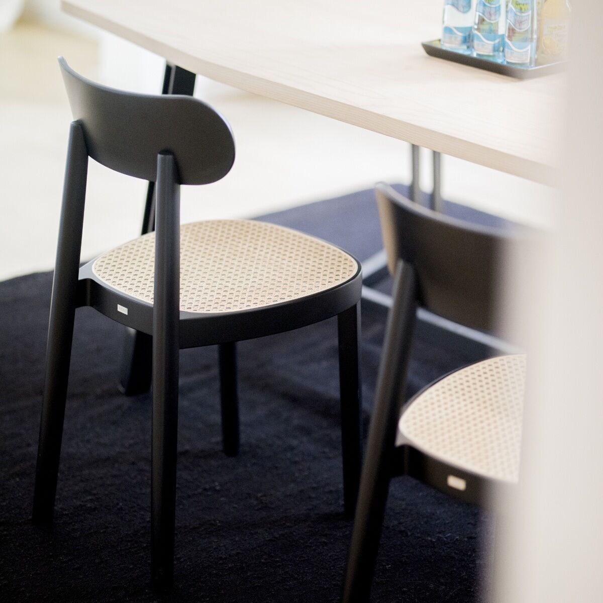 Ein moderner Stuhl von Thonet, Modell »118«, aus Holz mit schwarzer Rückenlehne und einem Sitz aus hellem Geflecht, steht vor einem hellen Tisch auf einer schwarzen Matte. Der Stuhl zeigt die typische hochwertige Verarbeitung von Thonet Möbeln.
