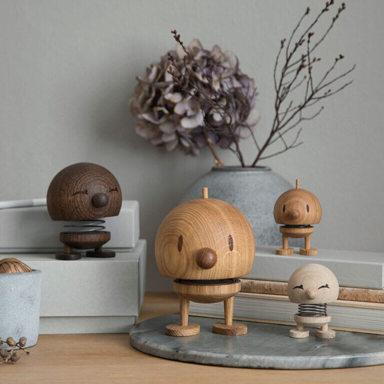 Ein Arrangement von Hoptimist Bumble Figuren aus Holz in verschiedenen Größen stehen auf einem Tisch vor einem grauen Hintergrund. Eine große Figur aus Eichenholz ist in der Mitte platziert, umgeben von weiteren Figuren in unterschiedlichen Holztönen. Im Hintergrund befinden sich getrocknete Blumen und Bücherstapel, die eine gemütliche und stilvolle Wohnatmosphäre schaffen.