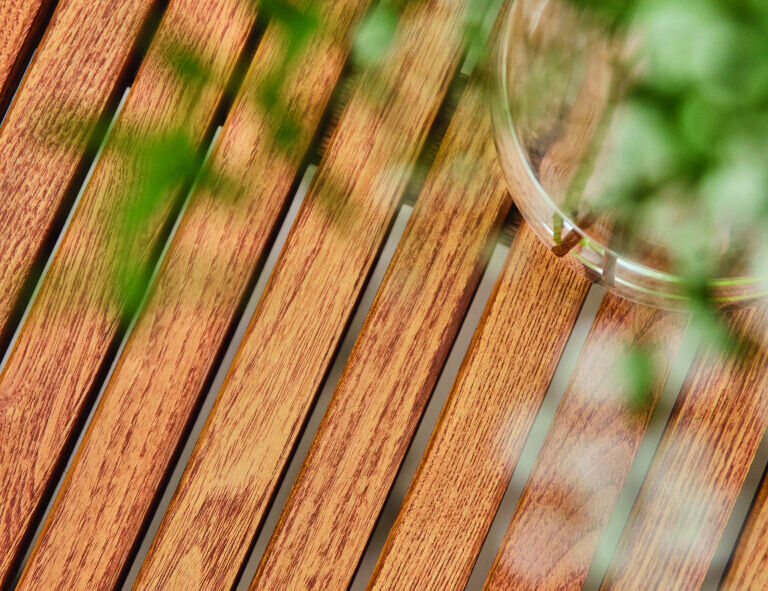 Nahaufnahme eines Holztisches mit grünen Pflanzen im Vordergrund und einem Glasbehälter auf der Tischoberfläche.