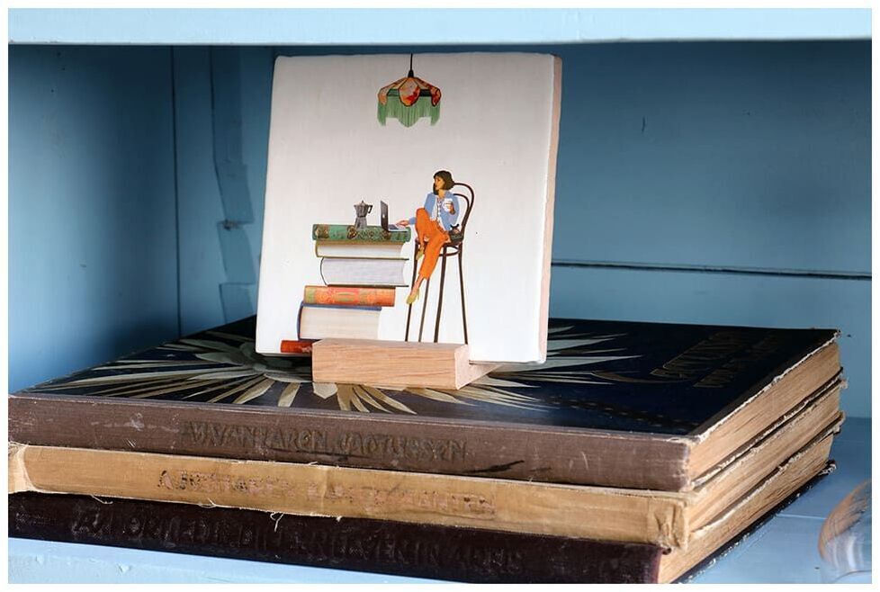 Keramikfliese mit der humorvollen Aufschrift »Alles unter Kontrolle« auf einem Holzständer, das auf einem Stapel Bücher in einem blauen Regal steht. Die Fliese zeigt eine Illustration von einem Mann, der in der Küche arbeitet.