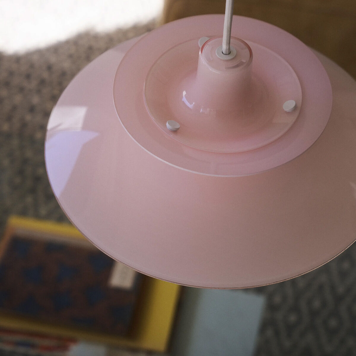 Hängeleuchte »PH 5 320« von Louis Poulsen mit glänzendem, pale-rosefarbenem Schirm aus Polymer, markantes dreistufiges Design, zentrische Aufhängung, moderner skandinavischer Stil. Unscharfer Hintergrund mit Teppich und Bücherstapel. Detailansicht von oben.