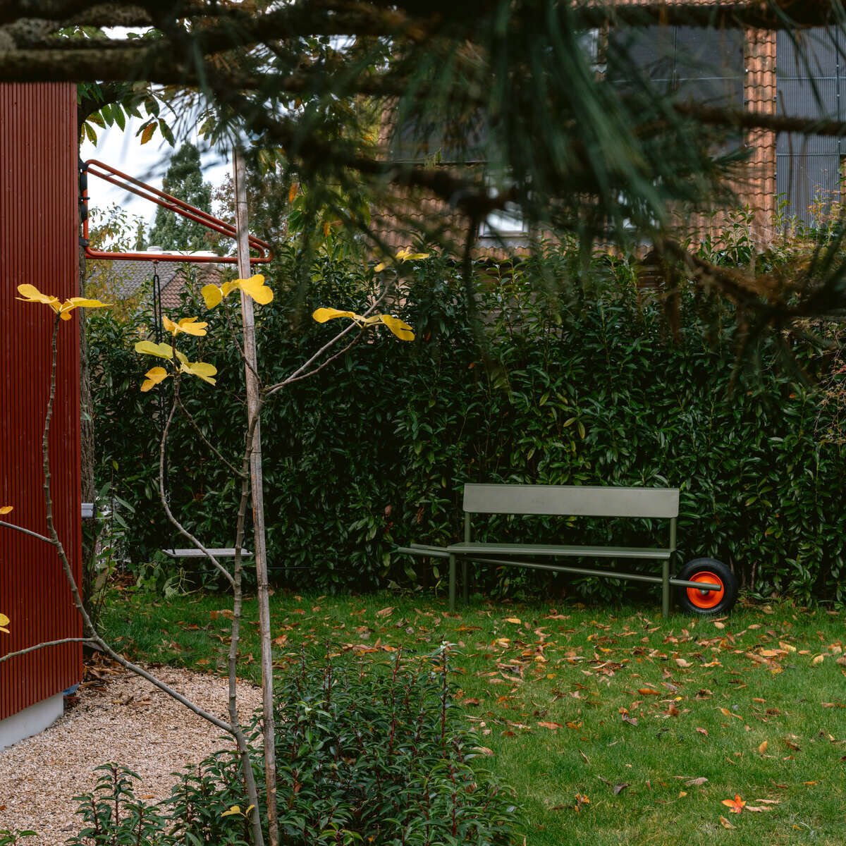 Im Bild steht die Schubkarrenbank »Wheelbench« von Weltevree an einer grünen Hecke in einem gepflegten Garten. Das Design ist modern, mit einem roten Rad für Mobilität. Rechts die Bank, links ein kleiner Baum, vorne Rasen, Büsche, im Hintergrund Klinkerhaus.