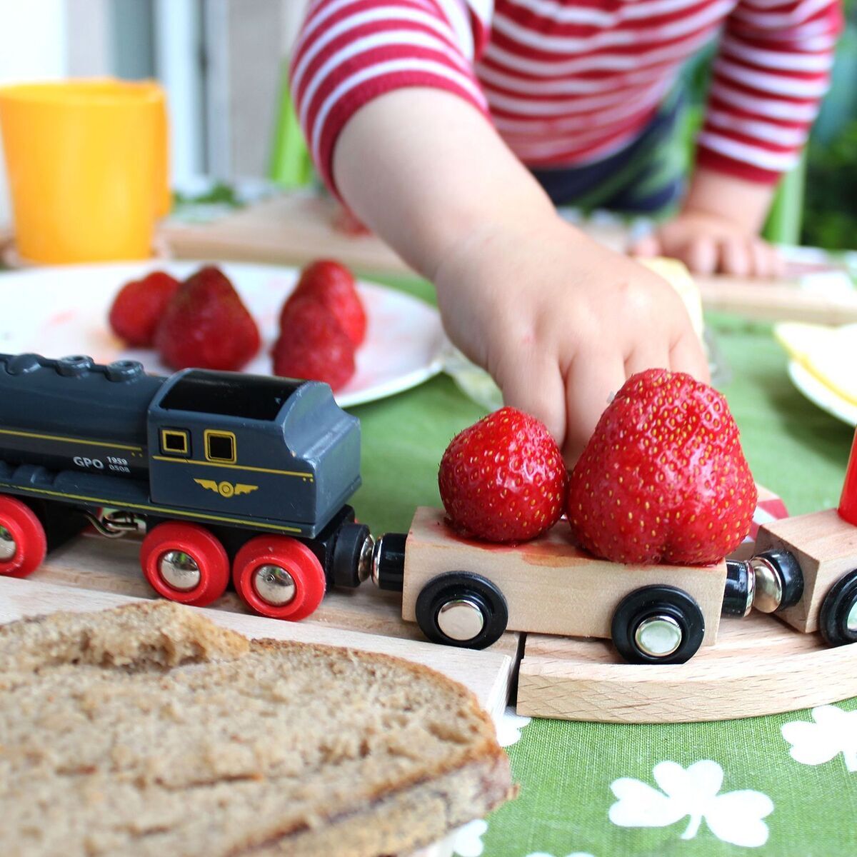 Ein Frühstücksbrett in Form einer Eisenbahn, darauf liegt Obst wie Erdbeeren und ein Spielzeugzug aus Holz. Im Hintergrund ist ein Kind mit einem gestreiften Pullover und weiteren Frühstücksgegenständen wie Brot und einer orangefarbenen Tasse zu sehen.