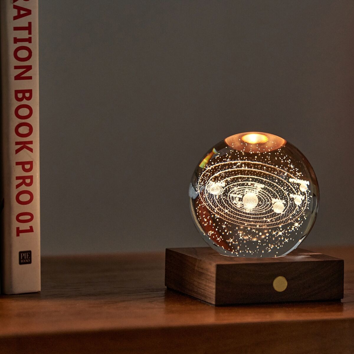 Eine stilvolle Lampe mit Kristallkugel im Sonnensystem-Design steht auf einem Sockel aus dunklem Walnussholz auf einem Holztisch. Das warme Licht erzeugt eine gemütliche Atmosphäre, daneben liegen eine Brille und ein Kissen auf einem Sofa. Im Hintergrund sind Bücher zu sehen.