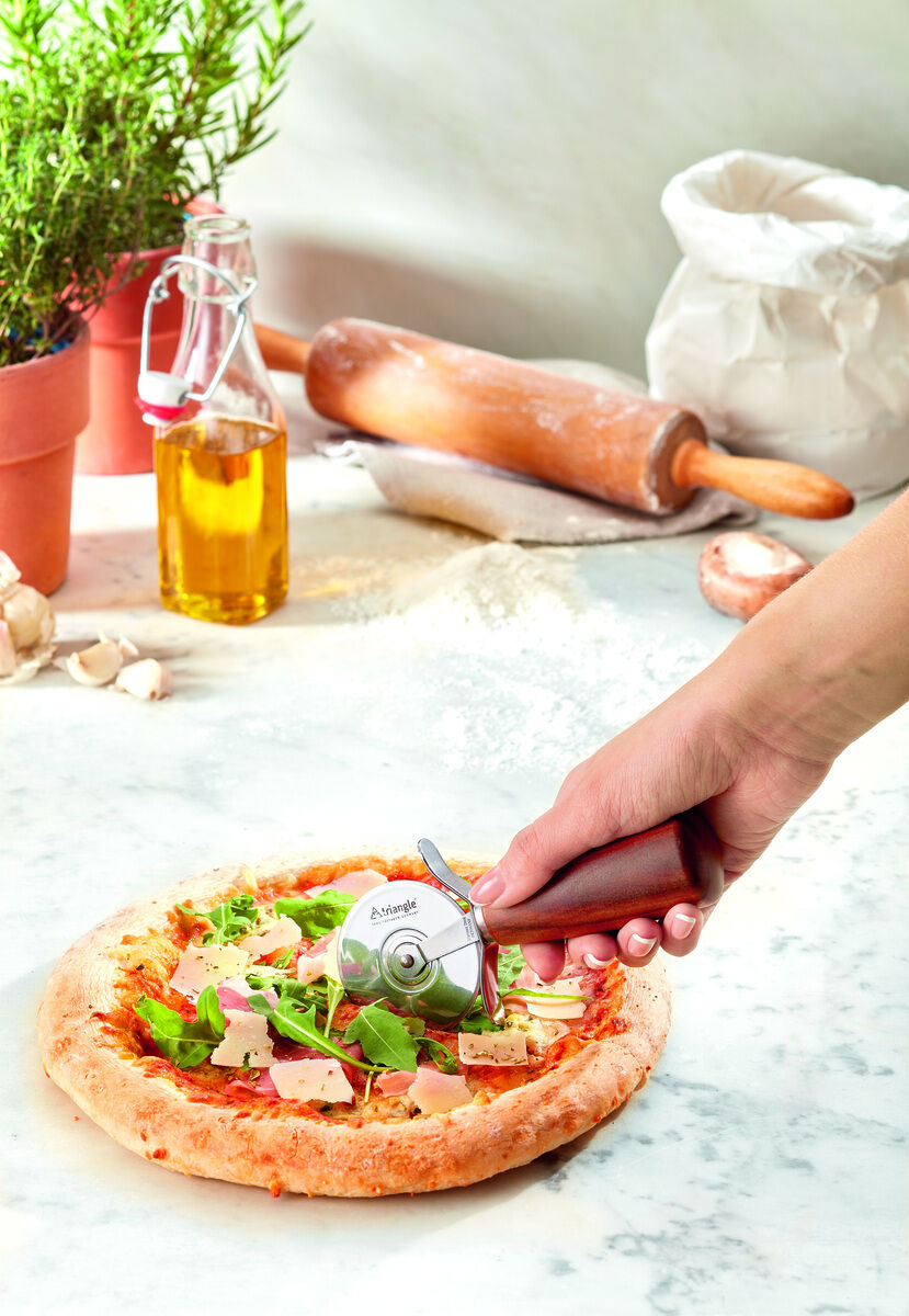 Eine Hand hält einen Pizzaschneider »Soul« aus Pflaumenholz über eine frisch gebackene Pizza. Im Hintergrund stehen Zutaten wie ein Olivenölflasche, Kräuter und ein Nudelholz auf einem weißen Marmortisch.