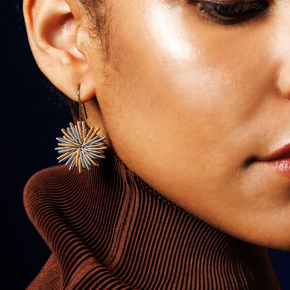 Nahaufnahme eines Ohrrings in stilisierter Blumenform aus Metall in Gold- und Silbertönen, getragen von einer Person mit braunem Rollkragenpullover. Das Design ist modern und auffällig, ideal für festliche oder besondere Anlässe.