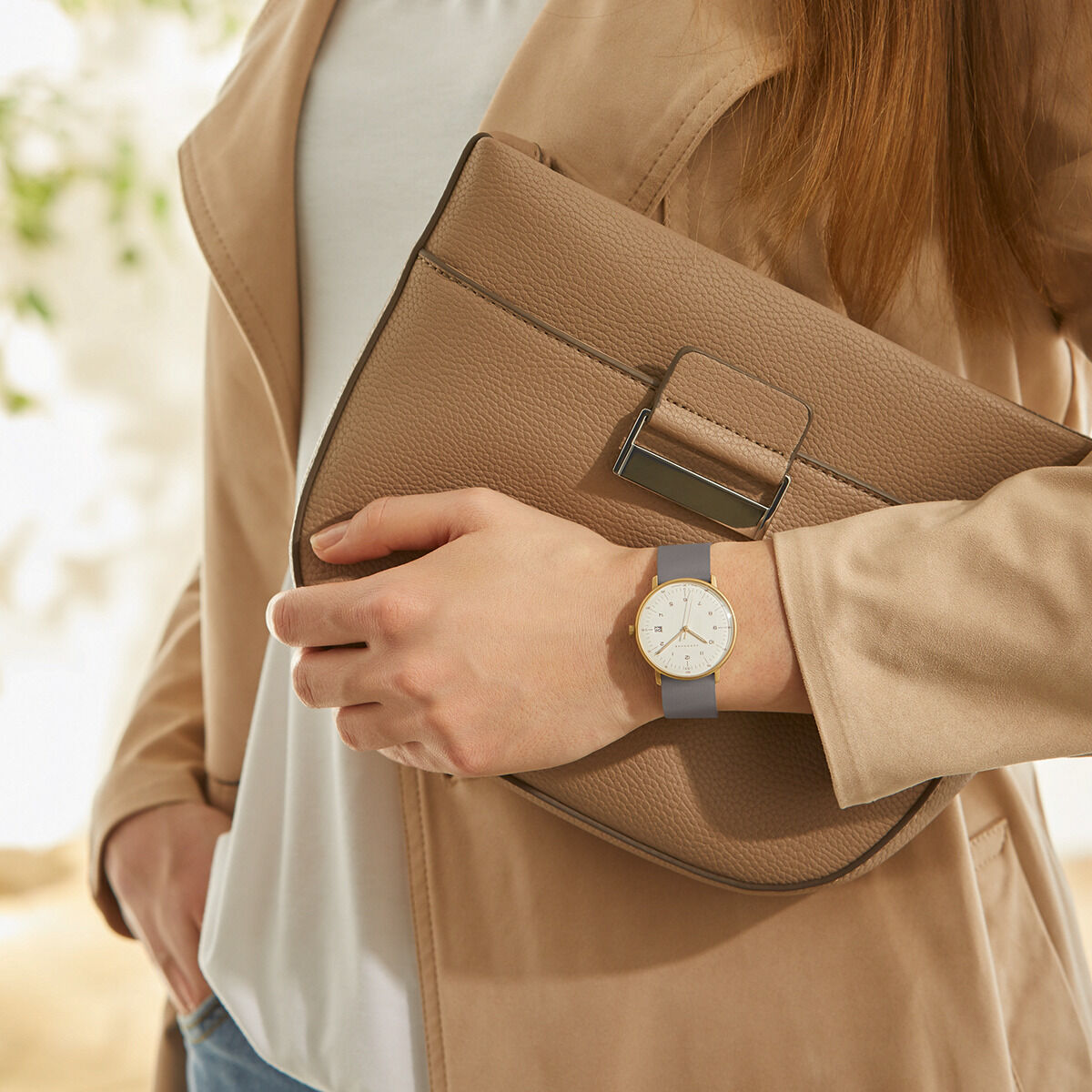 Eine elegante Damenarmbanduhr der ZEIT Sonderedition »max bill Damen«. Die Uhr hat ein schlichtes Design mit einem grauen Lederarmband und einem weißen Zifferblatt. Die Uhr wird an einem Handgelenk getragen und eine Frau hält eine beige Handtasche in der anderen Hand.
