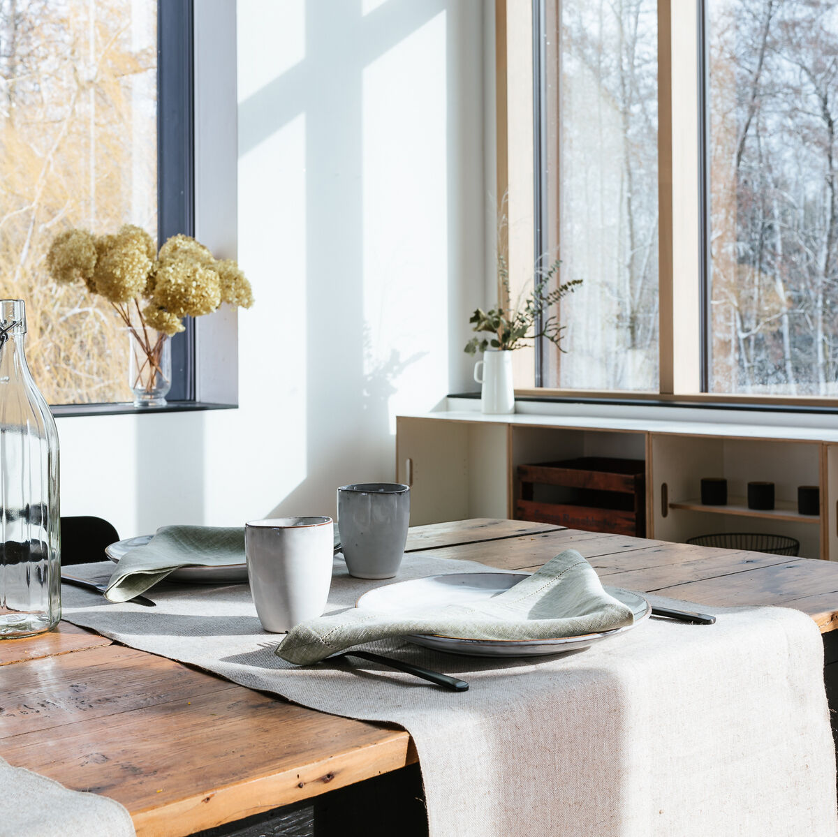 Ein gemütlich gedeckter Tisch in einen rustikalen Esszimmer. Ein Leinen-Tischläufer liegt auf einem Holztisch. Auf dem Tisch stehen weiße Keramik-Tassen und Teller, sowie leichte grüne Servietten. Im Hintergrund ist ein großes Fenster mit einer herbstlichen Baumlandschaft.