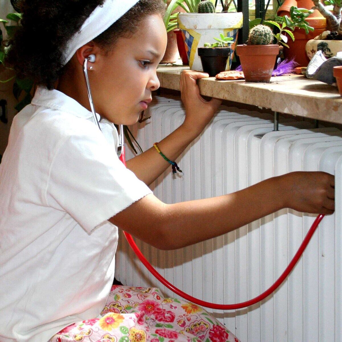 Ein Kind mit einem Stirnband benutzt einen roten Schlauch aus einem Experimentierkasten, um Geräusche von einem Heizkörper zu hören. Es gibt viele Topfpflanzen im Hintergrund auf der Fensterbank.