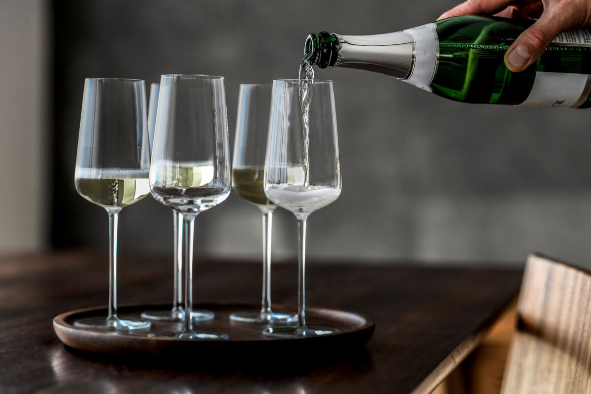 Eine Hand gießt Champagner in ein Set von vier schlanken Champagnergläsern auf einem runden Tablett. Die Szene ist elegant und modern gestaltet.