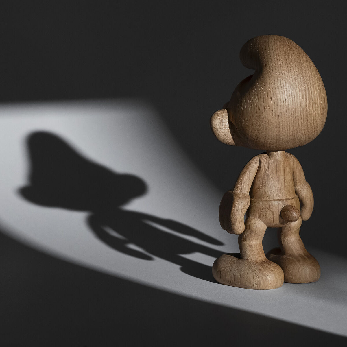 Holzfigur eines Schlümpfes steht unter Beleuchtung, die seinen Schatten auf den Boden wirft. Die Figur ist detailreich und in einem natürlichen Holzton gearbeitet.