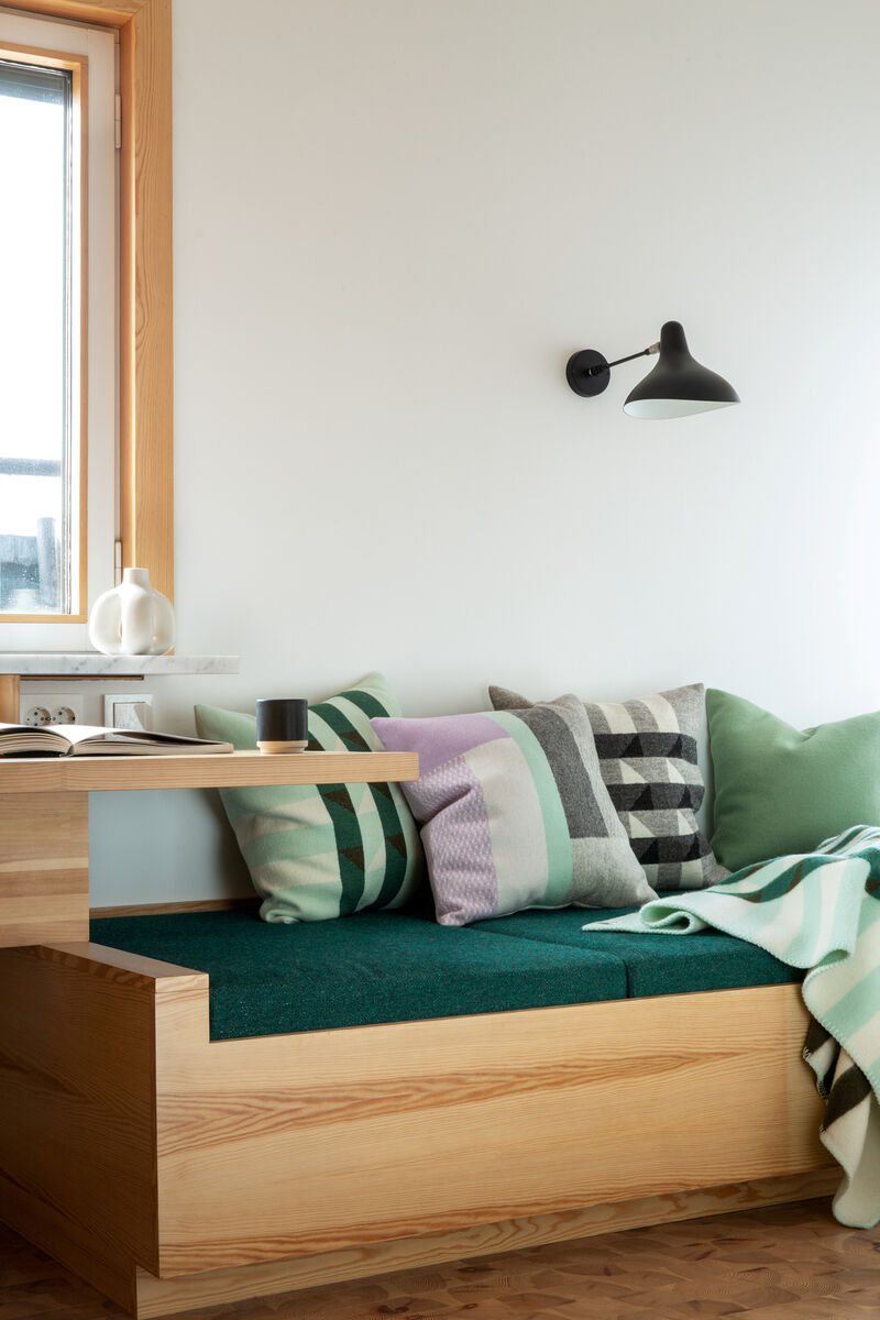 Gemütliche Wohnzimmer-Szene mit einer grünen Sofa-Ecke, dekoriert mit verschiedenen Kissen und einer grün-weiß gestreiften Lammwolldecke KVAM. Ein Holztisch und eine Lampe ergänzen den minimalistischen, skandinavischen Einrichtungsstil.