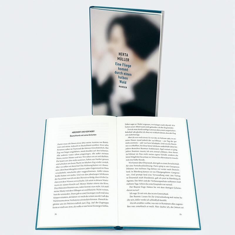 Das Bild zeigt das Buch 'Eine Fliege kommt durch einen halben Wald' von Herta Müller. Eine offene Seite des Buches ist im Vordergrund sichtbar, während das Buchcover im Hintergrund leicht unscharf dargestellt ist. Das Cover ist hauptsächlich in Weiß gehalten, mit einem verschwommenen Bild und dem Titel in schwarzen Buchstaben.