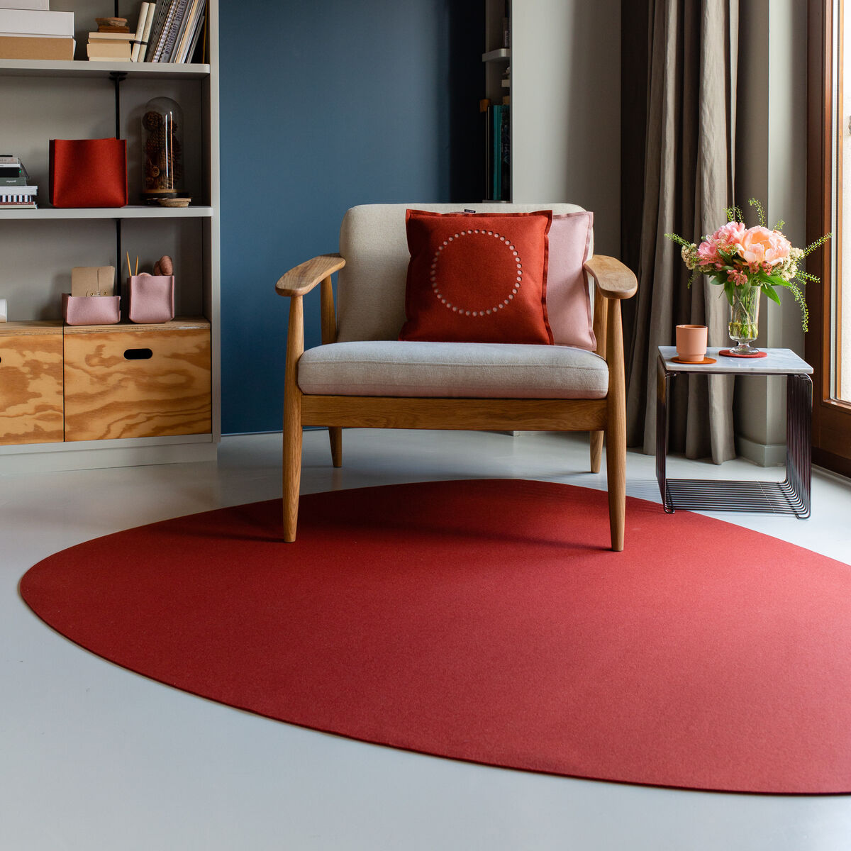Ein modernes Wohnzimmer mit einem runden, roten Teppich 'Stone', einem Holzstuhl mit beige gepolstertem Sitz und Rückenlehne, einem roten Kissen, einem Beistelltisch mit Blumenstrauß und einem Regal im Hintergrund.
