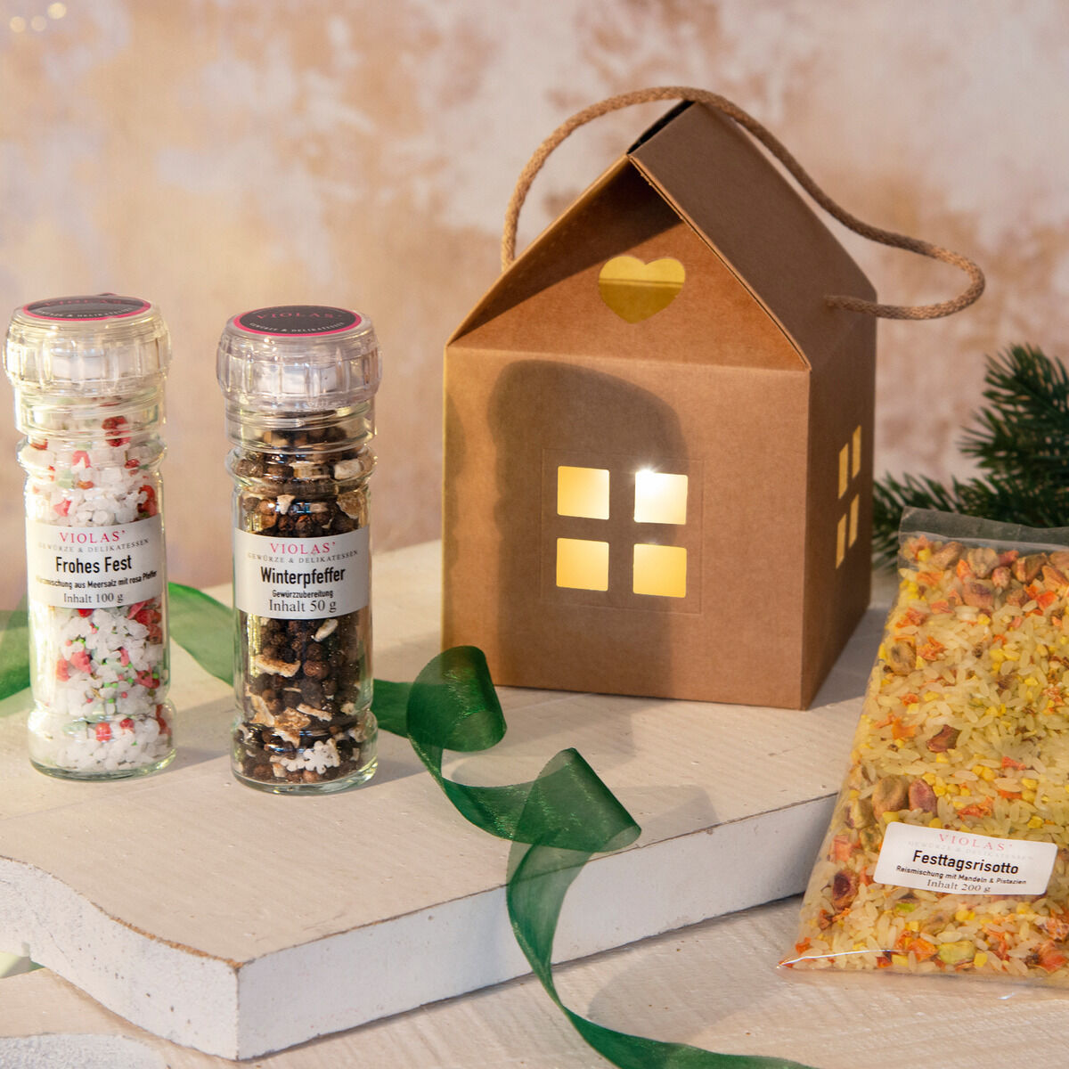 Geschenkset mit einem herzhaften Weihnachtshaus, zwei Gläsern von Gewürzen 'Fröhles Fest' und 'Winterpfeffer', sowie einer Packung Festtagsrisotto. Das Weihnachtshaus leuchtet warm. Ein grünes Band ergänzt die weihnachtliche Szenerie.