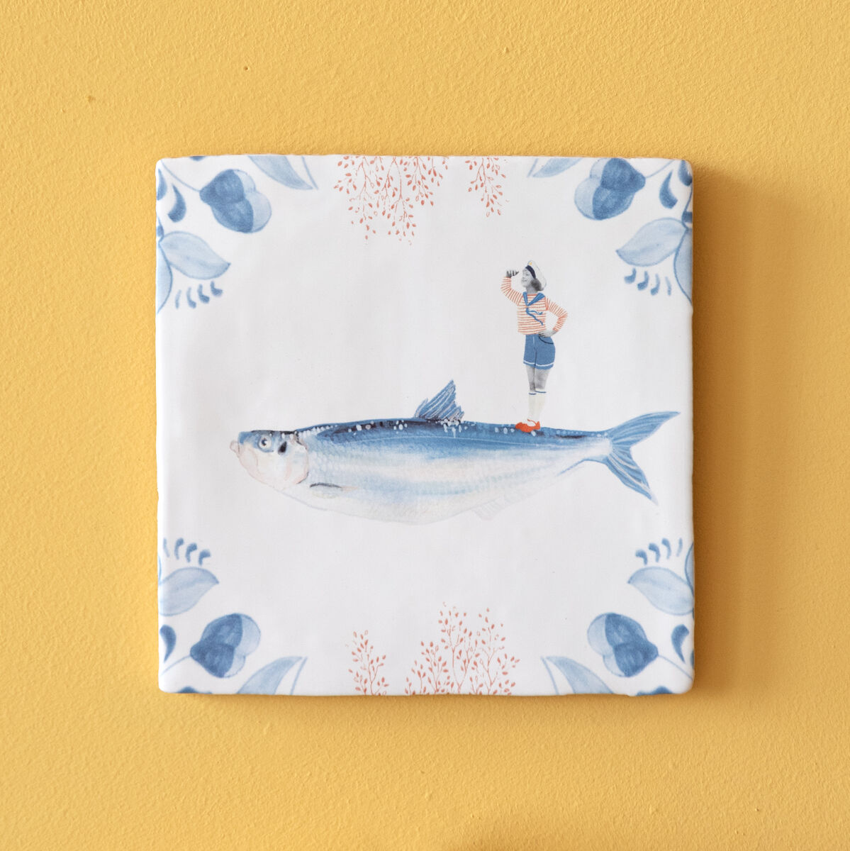 Keramikfliese »Blick nach vorn« mit einem Fischmotiv auf weißem Hintergrund, umgeben von blauen dekorativen Elementen. Befestigt an einer gelben Wand, darunter ein halber Teller in Blau und Terrakotta.