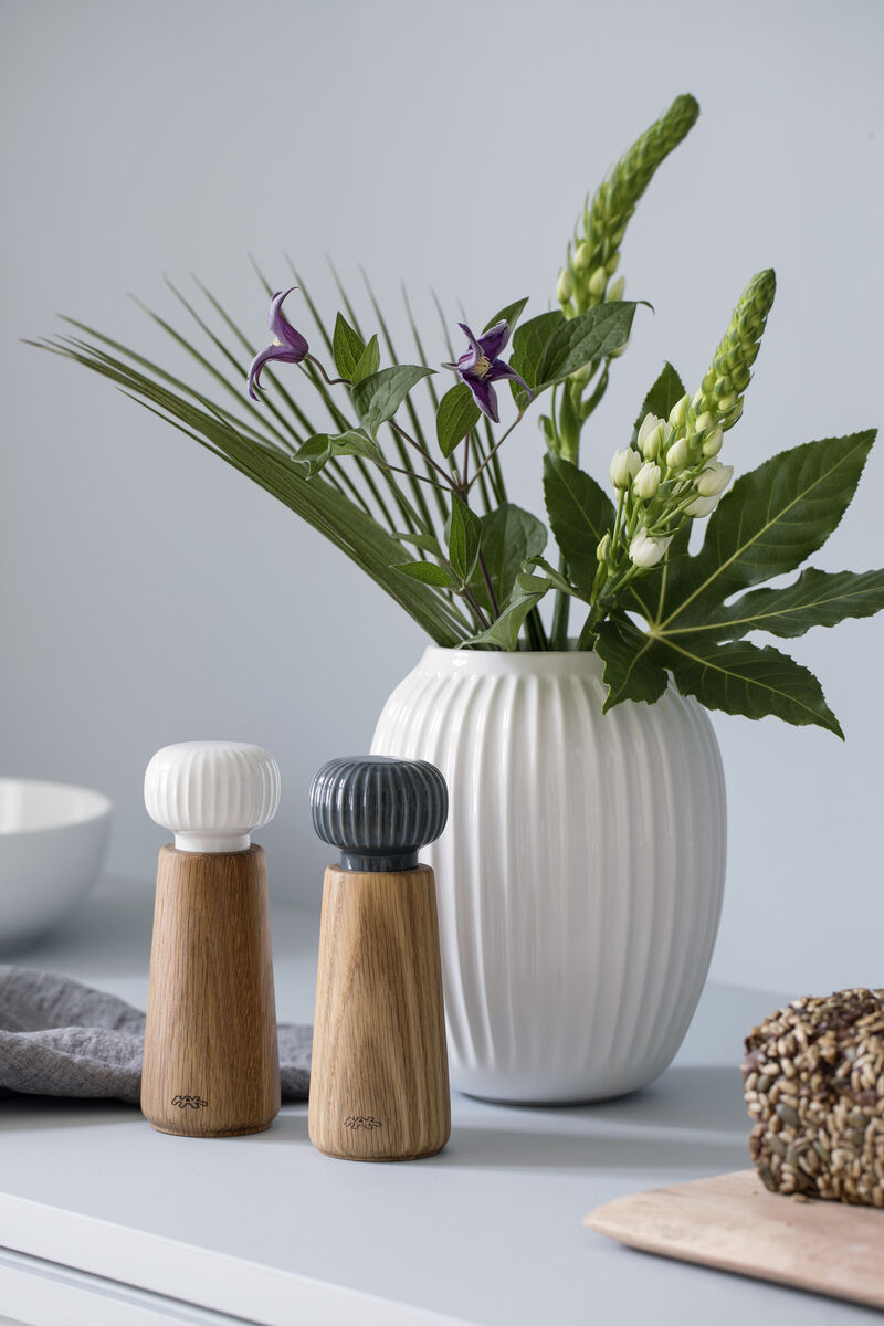 Zwei Gewürzmühlen der Serie »Hammershøi« aus Holz und Keramik stehen vor einer weißen Vase mit grünen und violetten Blumen auf einer weißen Oberfläche.