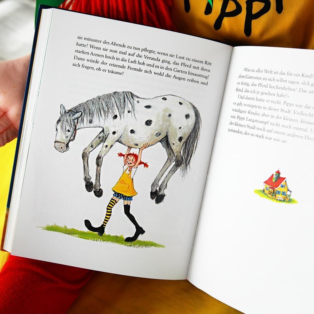 Ein geöffnetes Buch zeigt eine Illustration von Pippi Langstrumpf, die ein Pferd hochhebt. Der Hintergrund der Illustration ist weiß. Im unteren rechten Bereich des Bildes ist ein buntes Haus zu sehen. Eine Hand hält das Buch, und im Hintergrund ist ein Teil eines roten Kleidungsstücks zu sehen.