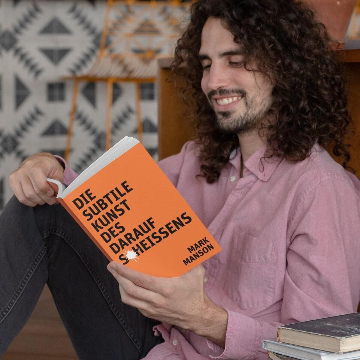 Ein junger Mann mit lockigem Haar sitzt auf dem Boden und liest ein Buch mit dem Titel 'Die subtile Kunst des darauf Scheißens' von Mark Manson. Das Buch hat ein orangefarbenes Cover mit schwarzem Text. Neben ihm liegt ein Stapel weiterer Bücher.