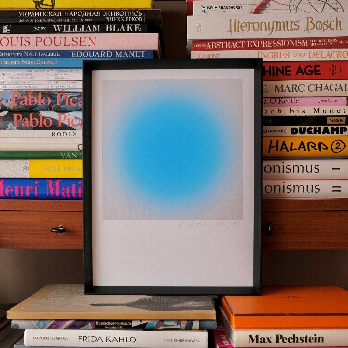 Der ZEIT-Sonderedition »Sorbet« Fine-Art-Print von Studio Anastasia Benko zeigt ein minimalistisches Kunstwerk in einem schwarzen Rahmen. Im Bildzentrum schwebt eine intensive, kreisförmige Blaunuance auf weißem Grund. Umgeben wird das Bild von farbenfrohen Kunst- und Designbüchern, auf und unter dem Print gestapelt.