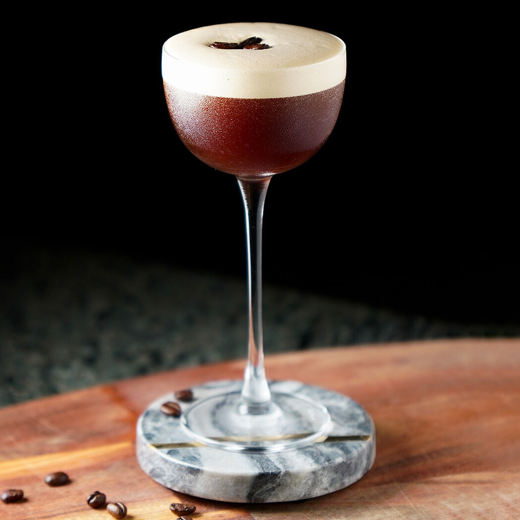 Ein mit Schaum bedeckter Kaffeelikör in einem edlen Cocktailglas mit Stiel, auf einem runden Marmorsockel mit einigen Kaffeebohnen, präsentiert auf einer hölzernen Oberfläche.