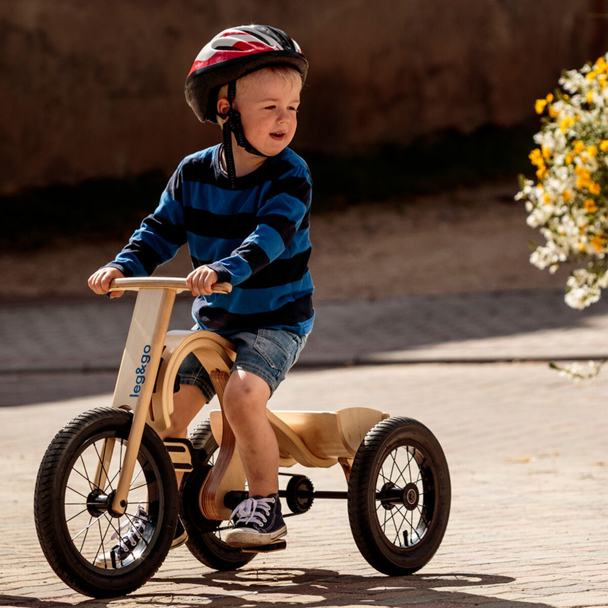 Kleines Kind mit blau-schwarz gestreiftem Pullover und Fahrradhelm fährt draußen bei Tageslicht ein Balance Bike mit Dreirad-Erweiterung aus Holz auf einem gepflasterten Weg. Im Hintergrund steht ein großer heller Blumentopf mit gelben und weißen Blüten.