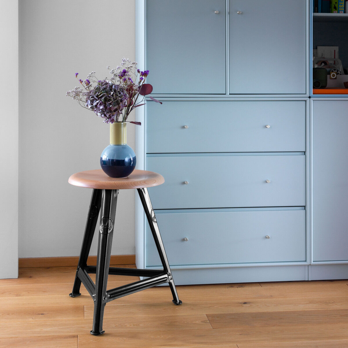 Rowac-Schemel aus Eiche in mattschwarz steht in einem modernen Raum mit hölzernem Boden und einer blauen Kommode im Hintergrund. Auf dem Schemel steht eine dekorierte Vase mit Blumen.
