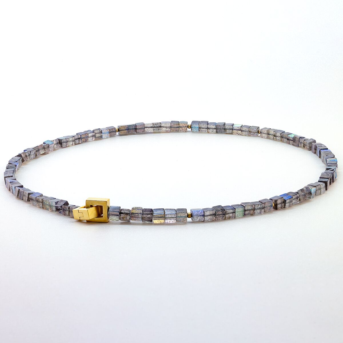 Ein Collier aus Labradorit-Würfeln mit feinem blaugrauem Schimmer, durchsetzt mit mattierten, goldplattierten Silberkugeln. Die Kette besitzt eine dezente, goldfarbene Patentschließe. Das Schmuckstück liegt kreisförmig vor einem weißen Hintergrund.