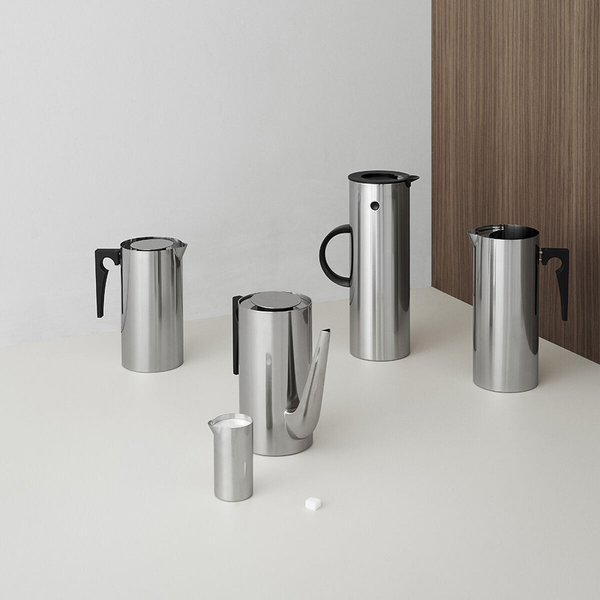Ein Set von Arne Jacobsen Design-Kaffeekannen aus Edelstahl, minimalistisch und modern gestaltet, auf einem weißen Tisch vor einer weißen und hölzernen Wand.