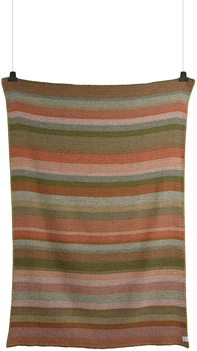 Gestreifte Lammwolldecke 'Fri' in Braun, Grün, Rosa und Beige, aufgehängt und fotografiert vor einem weißen Hintergrund.