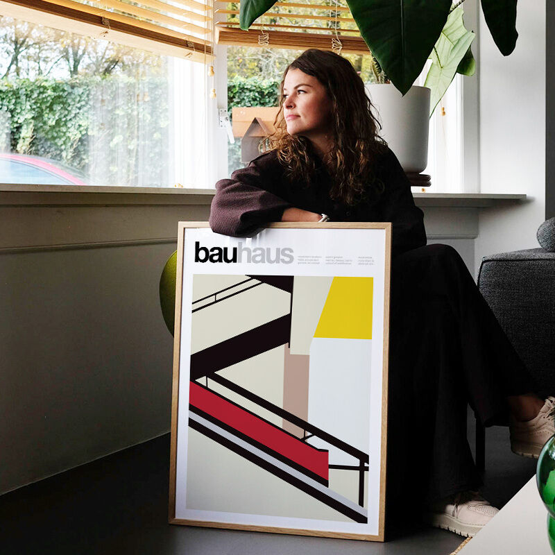 Frau sitzt neben einem gerahmten Poster mit dem Titel 'The Bauhaus Stairs', zeigt geometrische Formen in Schwarz, Rot, Gelb und Beige, typisch für den Bauhaus-Stil, an eine Wohnzimmerwand gelehnt.