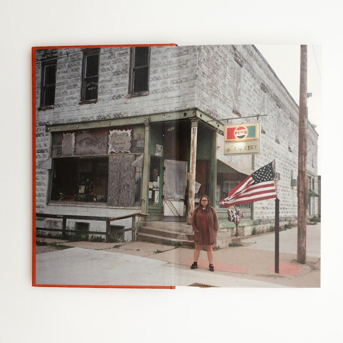 Foto von einem aufgeschlagenen Fotobuch mit einem Bild auf der rechten Seite, das eine Person vor einem alten, verlassenen Gebäude mit einer großen USA-Flagge zeigt. Die linke Seite des Buches ist leer sichtbar. Das Buch hat einen roten Einband.
