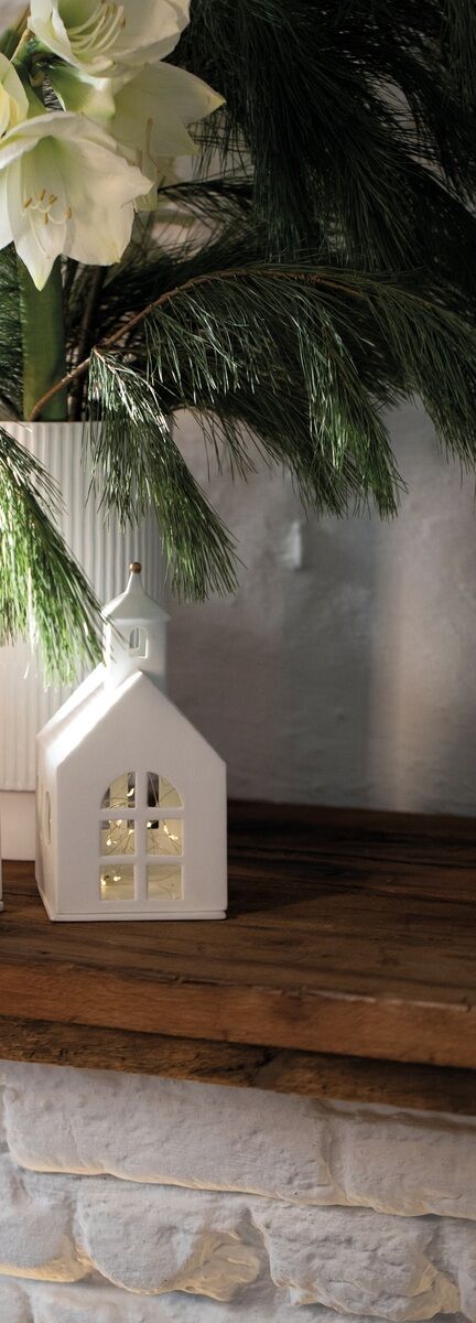 Eine weiße Kapelle als Lichthaus mit Kerze im Inneren, auf einem hölzernen Tisch, umgeben von Tannenzweigen und weißen Blumen in einer Vase.