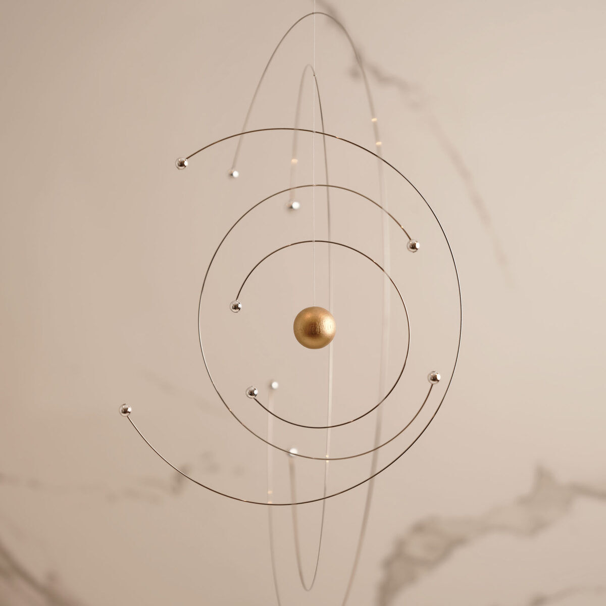 Ein modernes Mobile namens »Niels Bohr«, das physikalische Modell mit drei kreisförmigen Drähten darstellt, die sich um eine goldene Kugel im Zentrum drehen. Es ist in einem minimalistischen Stil gehalten und an einer unsichtbaren Schnur aufgehängt. Der Hintergrund ist ein einfacher beigefarbener Raum mit subtilen Marmorierungen.
