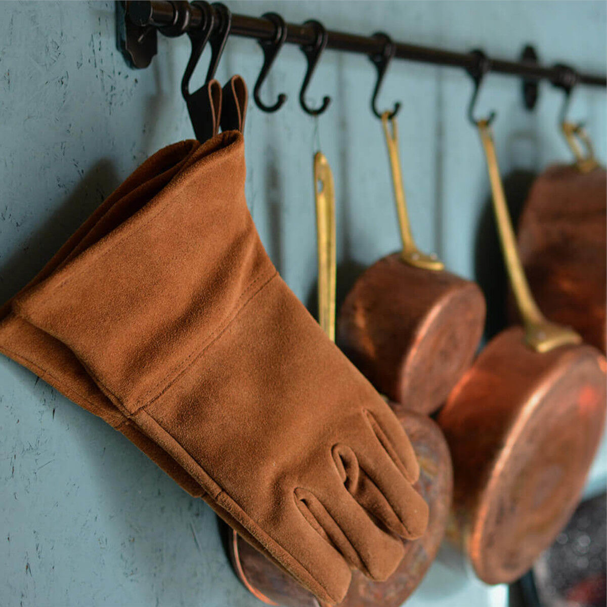 Ein Paar braune Rindleder-Handschuhe hängt an einer Hakenleiste neben mehreren Kupferpfannen in einer Küche.
