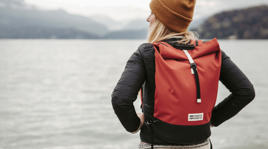 Person mit rotem Rucksack von Mero Mero blickt auf einen See mit Bergen im Hintergrund