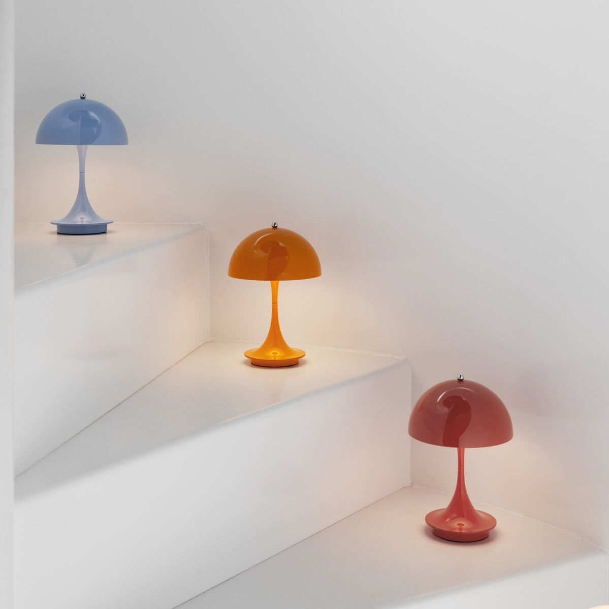 Ein Bild von drei Panthella 160 tragbaren Lampen in Blau, Orange und Rot, die auf weißen Treppen platziert sind. Die Lampen haben ein minimalistisches und modernes Design.