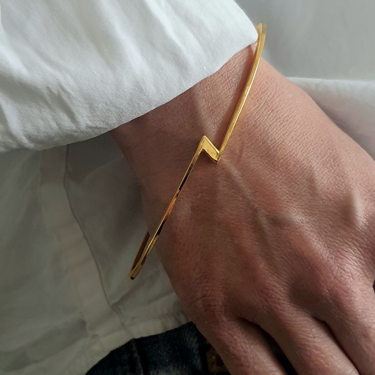 Ein stilvoller goldfarbener Armreif aus der ZEIT-Jubiläumsedition wird an einem Handgelenk getragen. Der Armreif hat ein modernes, minimalistisches Design. Er wird mit einer weißen Bluse und einer blauen Jeans kombiniert.