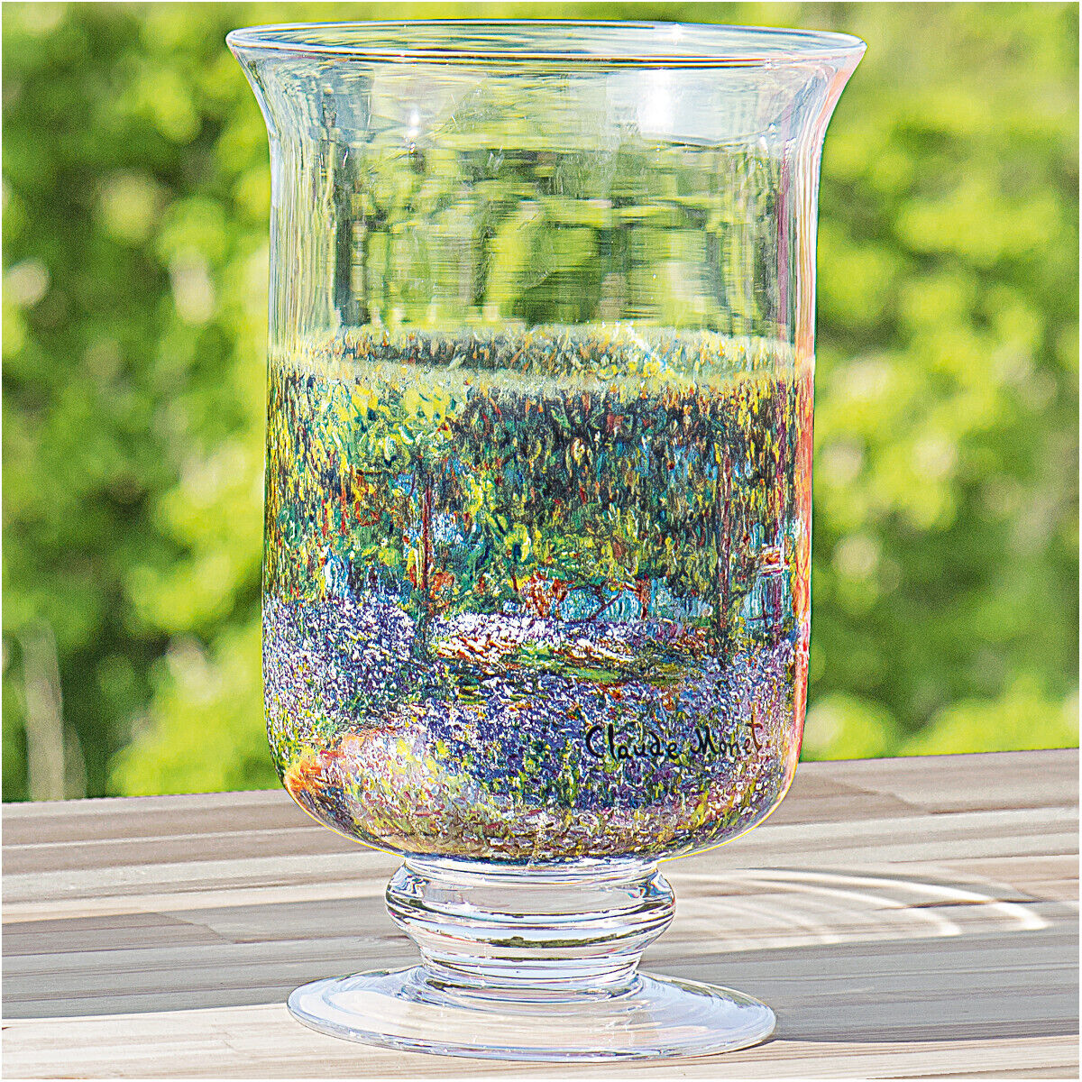 Ein handbemaltes Windlicht oder Vase aus Glas mit einem impressionistischen Gemälde von Claude Monet, das eine bunte Natur- und Landschaftsszene darstellt. Das Glasobjekt steht auf einem Holztisch mit einem verschwommenen grünen Hintergrund.