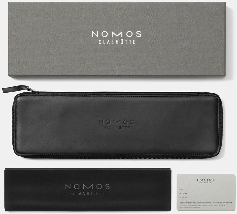 Abgebildet ist die edle NOMOS Glashütte Verpackung: eine graue Box mit weißem Logo, ein elegantes schwarzes Etui mit Reißverschluss, ein passendes Lederetui sowie eine Garantiekarte. Das Set wirkt hochwertig, minimalistisch und für luxuriöse Uhren geeignet.